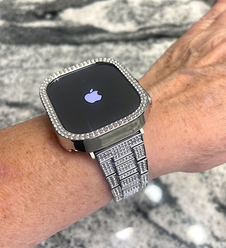 Op de afbeelding: Een zilveren Apple Watch met een band en bezel bezet met diamanten. De wijzerplaat van het horloge is zwart en toont het Apple-logo.