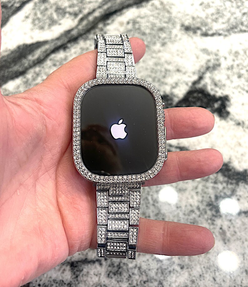 Op de afbeelding: Een zilveren Apple Watch met een band en bezel bezet met diamanten. De wijzerplaat van het horloge is zwart en toont het Apple-logo.