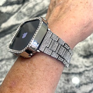 Op de afbeelding: Een zilveren Apple Watch met een band en bezel bezet met diamanten. De wijzerplaat van het horloge is zwart en heeft het Apple-logo.