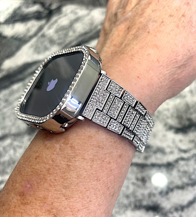 Op de afbeelding: Een zilveren Apple Watch met een band en bezel bezet met diamanten. De wijzerplaat van het horloge is zwart en heeft het Apple-logo.