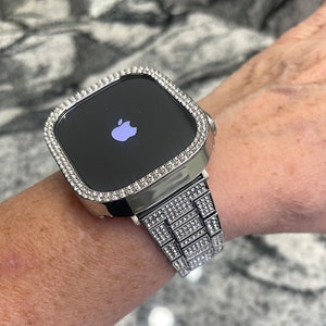 Op de afbeelding: Een zilveren Apple Watch met een band en bezel bezet met diamanten. De wijzerplaat van het horloge is zwart en toont het Apple-logo.