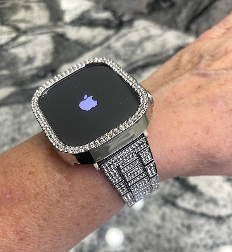Op de afbeelding: Een zilveren Apple Watch met een band en bezel bezet met diamanten. De wijzerplaat van het horloge is zwart en toont het Apple-logo.