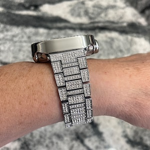 Op de afbeelding: Een zilveren horloge met een band en wijzerplaat bezet met diamanten. Het horloge zit om de pols van een persoon.