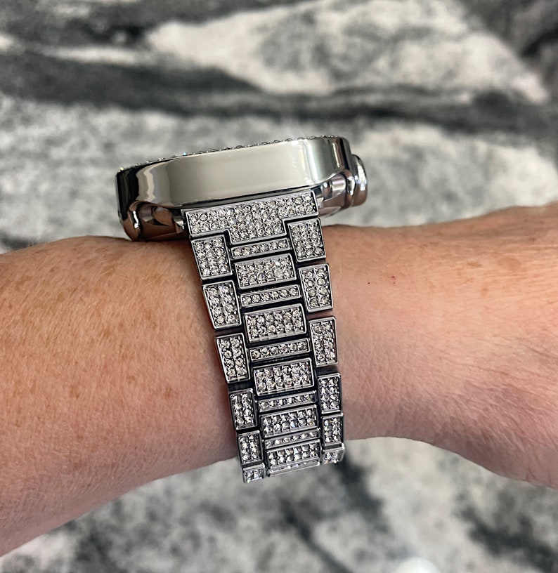 Op de afbeelding: Een zilveren horloge met een band en wijzerplaat bezet met diamanten. Het horloge zit om de pols van een persoon.