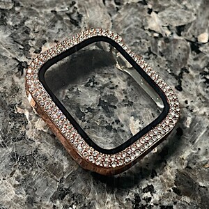 Puede incluir: Una carcasa de Apple Watch de oro rosa con un protector de pantalla transparente y un bisel incrustado con cristales que parecen diamantes.