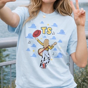 Puede incluir: Una camiseta azul claro con un gr&aacute;fico de una persona de cabello rubio que viste una camisa de cuadros amarillos y pantalones con estampado de vaca, sosteniendo una guitarra y un sombrero rojo. El dise&ntilde;o incluye las letras TS, notas y nubes.