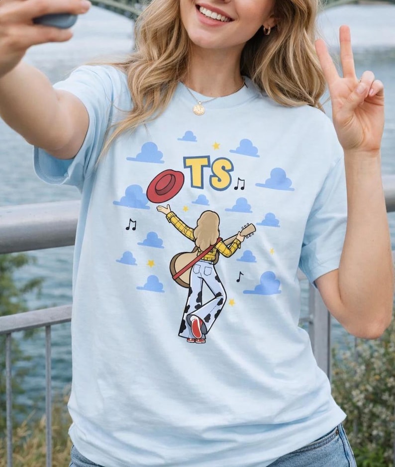 Puede incluir: Una camiseta azul claro con un gr&aacute;fico de una persona de cabello rubio que viste una camisa de cuadros amarillos y pantalones con estampado de vaca, sosteniendo una guitarra y un sombrero rojo. El dise&ntilde;o incluye las letras TS, notas y nubes.