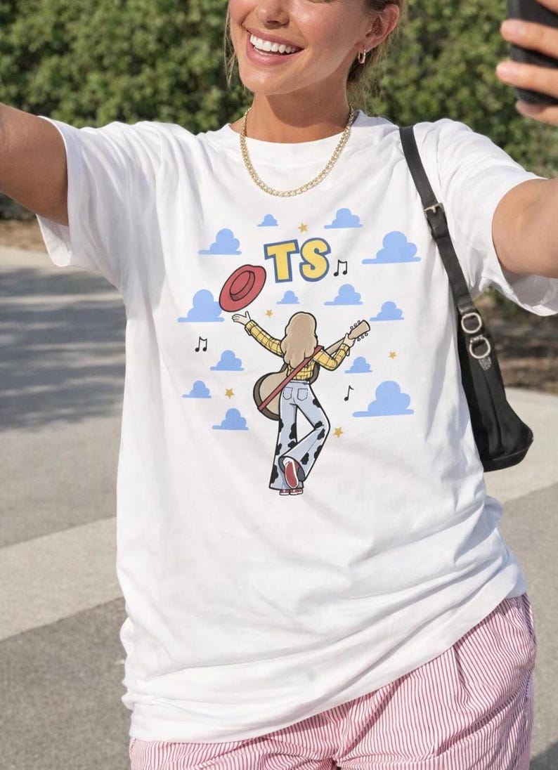 Puede incluir: Una camiseta blanca con un gr&aacute;fico de una persona de cabello rubio que viste una camisa de cuadros amarillos y pantalones con estampado de vaca, sosteniendo una guitarra y un sombrero rojo. El dise&ntilde;o incluye nubes azules, estrellas amarillas, notas musicales y las letras TS.