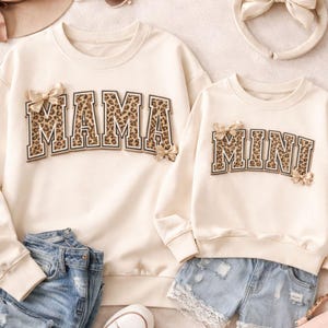 May include: Cream-colored sweatshirts, one adult and one child size, with the words "MAMA" and "MINI" in leopard print lettering. Each sweatshirt has small bow accents. Denim shorts and jeans are visible.