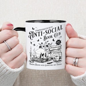 May include: White ceramic mug with a black handle and rim, featuring a gothic design. The design includes a skull, books, candles, and the text "UNAPOLOGETICALLY ANTI-SOCIAL BOOK CLUB". The phrase "MAKING FRIENDS WITH CHARACTERS, NOT PEOPLE" is also printed on the mug.