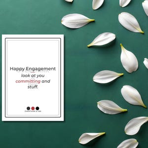Może przedstawiać: Biała karta z napisem "Happy Engagement" i słowami "look at you committing and stuff." na ciemnozielonym tle. Białe płatki lilii i kwiat lilii są rozrzucone wokół karty.