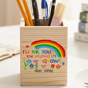 Könnte beinhalten: Ein quadratischer Stiftehalter aus Holz mit einem bunten Aufdruck aus Regenbogen, Blumen und einem Herz. Der Text lautet: THANK YOU FOR HELPING US GROW! FROM, SOPHIA. Der Halter enthält mehrere Bleistifte, Stifte und einen Textmarker.