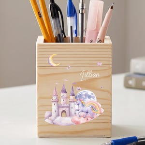 May include: A wooden pencil holder with a whimsical castle and unicorn design. The holder contains pencils, pens, and a highlighter. The name "Jillian" is written on the front. A red apple and books are in the background.