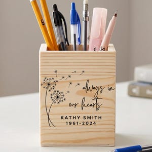 Op de afbeelding: Een houten pennenbakje met de tekst "always in our hearts" en een paardenbloemontwerp. De houder bevat potloden, pennen en een roze markeerstift. De tekst bevat een naam en data.