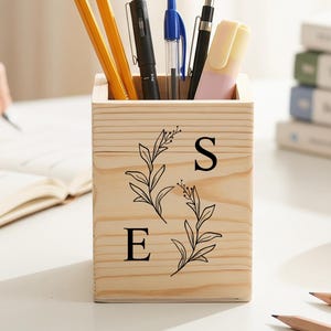 Può includere: Un portapenne in legno con matite, penne e un evidenziatore. Il portapenne è decorato con un motivo floreale e le lettere "S" ed "E". Sono visibili anche un libro aperto, una pila di libri e una mela.
