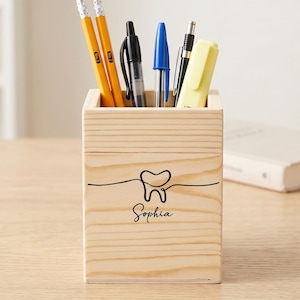 Peut inclure: Un porte-crayon carré en bois clair orné d'un dessin au trait noir représentant une dent et le prénom Sophia écrit en lettres cursives. Le pot contient deux crayons jaunes, un stylo noir, un stylo bleu et un surligneur jaune.