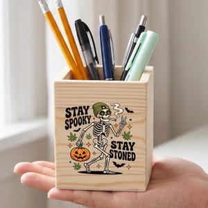May include: A square wooden pencil holder featuring a graphic of a skeleton wearing a green beanie, holding a pumpkin, and making a peace sign. The text "STAY SPOOKY" and "STAY STONED" is printed in black, accompanied by small bats and leaf icons.