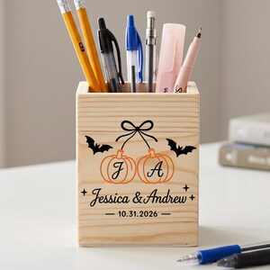 May include: A square light-colored wooden pencil holder featuring a black graphic of two pumpkins hanging from a bow, two bats, and the text "J A", "Jessica & Andrew", and "10.31.2026". The holder contains various pencils, pens, and markers.