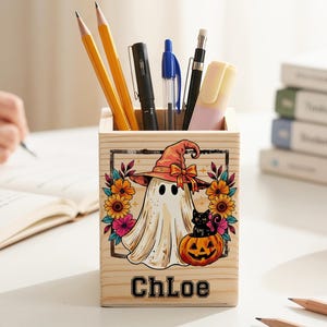 May include: A square wooden pencil holder featuring a printed graphic of a ghost wearing a witch hat, a black cat sitting in a pumpkin, and colorful flowers. The name Chloe is printed in black block letters at the bottom of the wooden container.