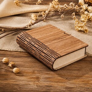 May include: A rectangular wooden box with a hinged lid featuring a smooth grain finish on the top surface and a decorative, laser-cut slatted pattern along the curved side edge. The box has a light brown tone and a flat, solid base construction.
