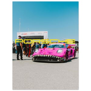 May include: A bright pink Porsche race car with a shark-mouth design on the front. The car is parked on a paved surface with people and a banner in the background. The word "PORSCHE" is displayed in yellow.