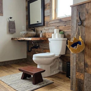 May include: A yellow felt crown with a blue interior lining hangs from a black metal hook attached to a vertical reclaimed wood panel. The crown features a scalloped top edge and is positioned in a bathroom setting with a toilet and stool.