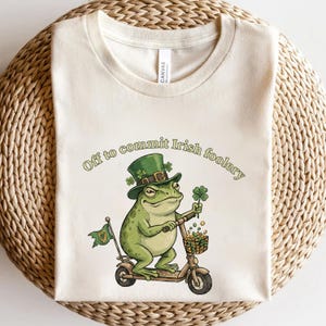 以下が含まれることがあります： クリーム色のTシャツ。緑色のカエルがスクーターに乗り、緑色のシルクハットをかぶり、四つ葉のクローバーを持っています。「Off to commit Irish foolery」の文字がカエルの上にアーチ状に書かれています。