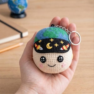 May include: A handmade crochet keychain featuring a smiling face with a blue and green globe hat and a black eye mask with stars, a crescent moon, and rockets. The keychain is attached to a silver metal ring.