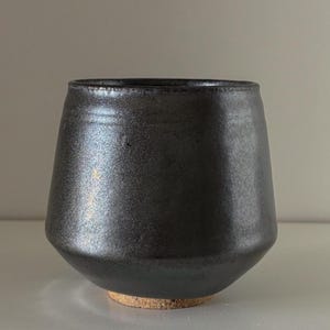 May include: A black ceramic tumbler with a wide mouth and a slightly tapered body. The exterior has a metallic sheen, and the base rests on a natural, unglazed ceramic ring. The tumbler is designed for beverages.