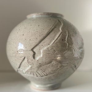 May include: A speckled, round ceramic vase in a neutral color. The vase features a textured, carved design on its surface. The rim and base are a lighter shade, creating a subtle contrast. A decorative piece for home decor.