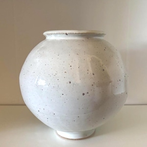 Könnte beinhalten: Eine kugelförmige Keramikvase mit glänzender weißer Glasur und dunkelbraunen Sprenkeln. Die Vase hat einen schmalen, leicht ausgestellten Rand an der Oberseite und einen kleinen, kreisförmigen Boden, was ihre abgerundete, bauchige Körperform unterstreicht.