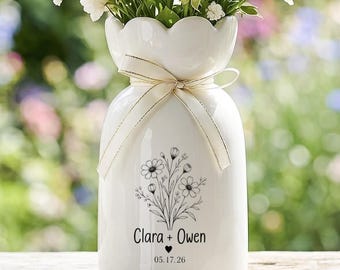 Può includere: Un vaso in ceramica bianca con bordo smerlato e un nastro color crema legato attorno al collo. La parte anteriore presenta un disegno floreale nero, il testo Clara + Owen, un piccolo cuore e la data 05.17.26, riempito di fiori gialli e bianchi.