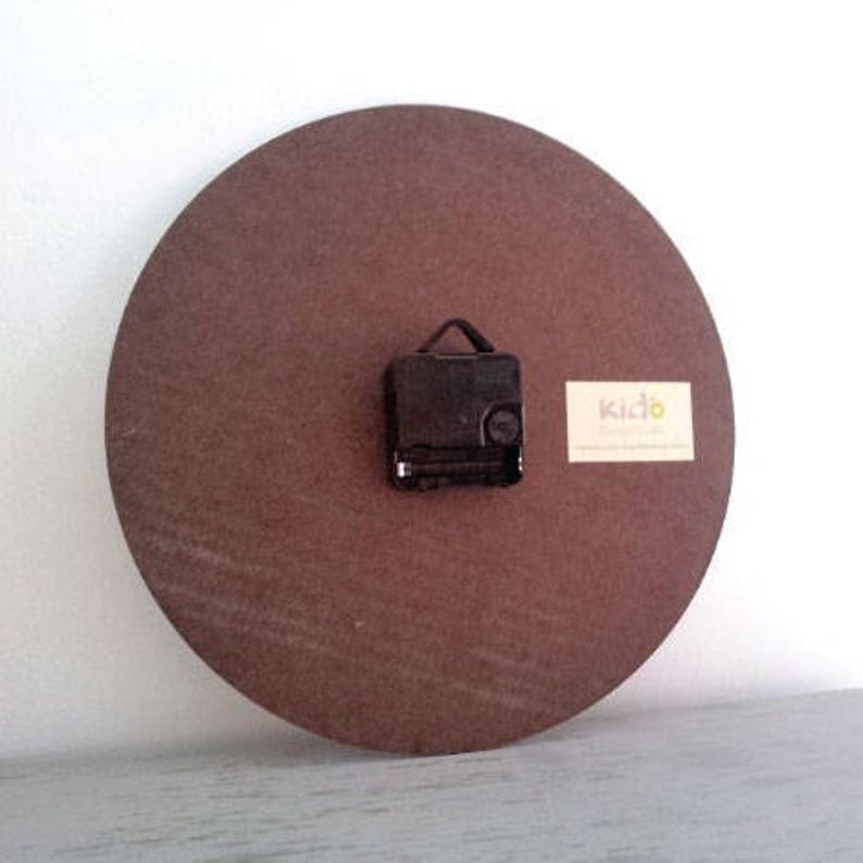 May include: The back of a brown wooden clock with a black clock mechanism and a white label with the text "Kido" printed on it.