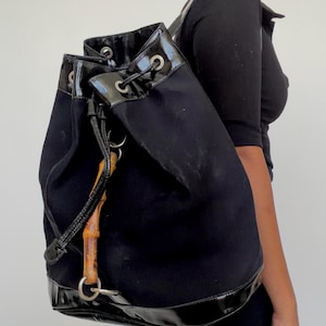 Könnte beinhalten: Schwarzer Canvas-Rucksack mit Kordelzugverschluss und glänzendem schwarzen Besatz. Die Tasche hat einen Bambusgriff und silberfarbene Hardware. Der Rucksack ist in einer Eimerform gestaltet und wird über der Schulter getragen.