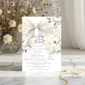 May include: A bridal shower invitation featuring a large, elegant bow and white floral accents. The invitation includes the text "she's tying the knot" and event details. The design is soft and romantic, with a focus on a wedding theme.