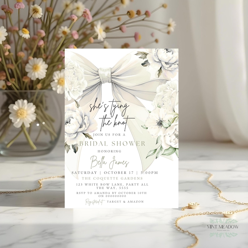 May include: A bridal shower invitation featuring a large, elegant bow and white floral accents. The invitation includes the text "she's tying the knot" and event details. The design is soft and romantic, with a focus on a wedding theme.