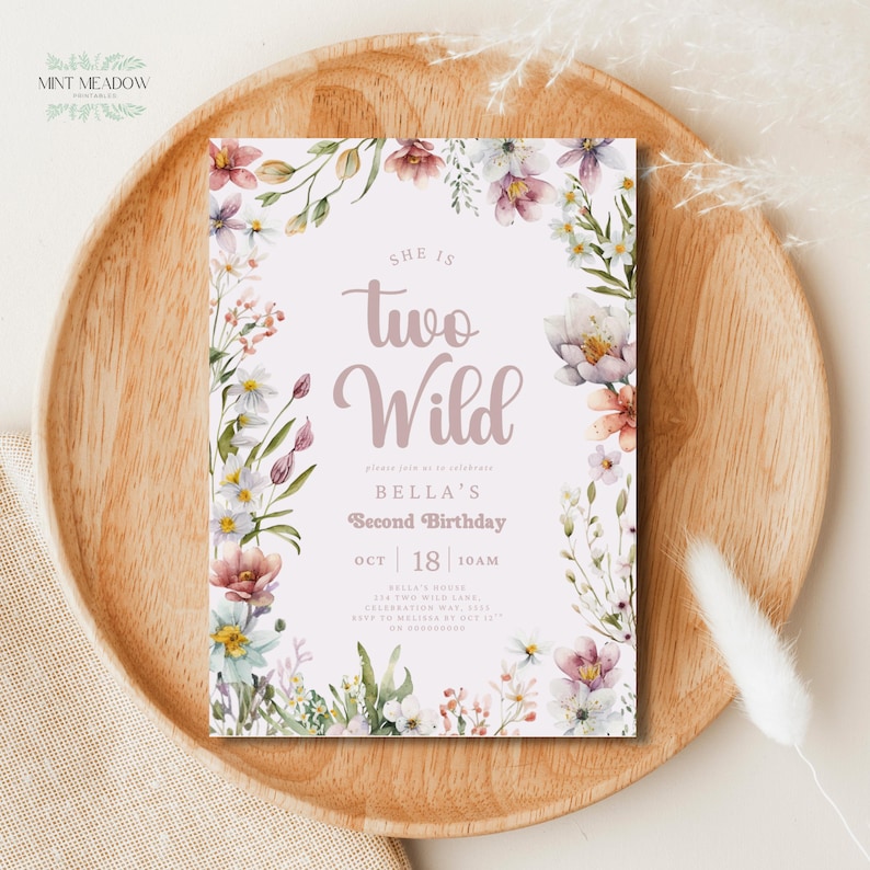 May include: A floral-themed birthday invitation with the text "She is Two Wild" in a soft, cursive font. The invitation is surrounded by watercolor flowers in shades of pink, white, and green. The invitation is on a wooden tray.