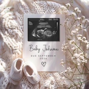 May include: A pregnancy announcement card featuring a sonogram image, the name "Baby Johnson", and the due date "September 2026". Two small white knitted baby booties are placed below the card. The card is on a white knitted blanket, with white flowers.