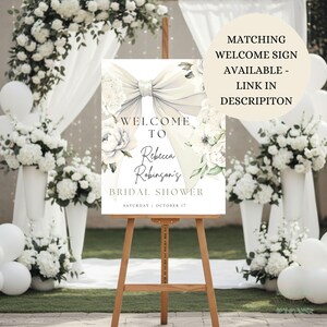 May include: A wooden easel displays a welcome sign for Rebecca Robinson's bridal shower. The sign features a large bow and floral accents, with the text "Welcome To Rebecca Robinson's Bridal Shower" and the date "Saturday | October 17".