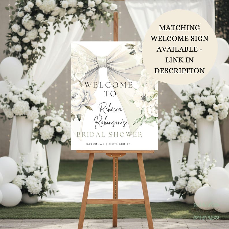 May include: A wooden easel displays a welcome sign for Rebecca Robinson's bridal shower. The sign features a large bow and floral accents, with the text "Welcome To Rebecca Robinson's Bridal Shower" and the date "Saturday | October 17".