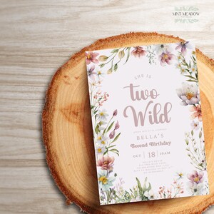 May include: A floral-themed birthday invitation with the text "She is Two Wild" in a cursive font. The invitation is surrounded by watercolor flowers in shades of pink, purple, and white. The invitation is on a wooden surface.