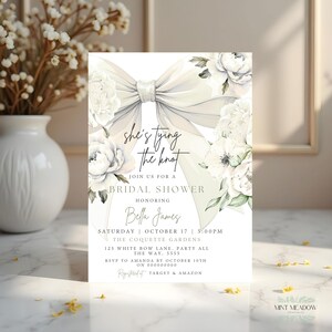 May include: Bridal shower invitation with a large, elegant bow and floral accents. The invitation features the text "she's tying the knot" and event details, including the date, time, and location. The design is soft and romantic.