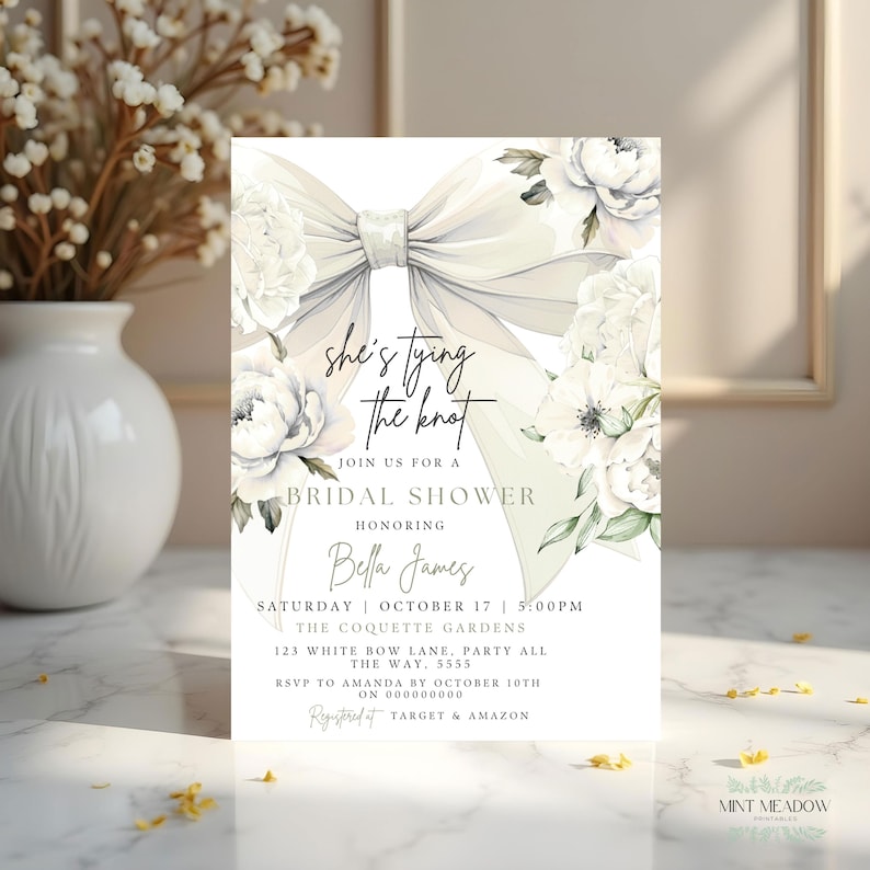 May include: Bridal shower invitation with a large, elegant bow and floral accents. The invitation features the text "she's tying the knot" and event details, including the date, time, and location. The design is soft and romantic.