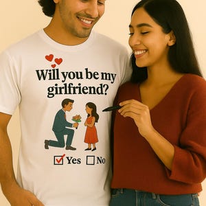 Puede incluir: Una camiseta blanca con el texto "¿Quieres ser mi novia?" y un gráfico de un hombre proponiendo matrimonio a una mujer. La camiseta también tiene una casilla de verificación "Sí". Una mujer con un suéter rojo y jeans azules está al lado del hombre.