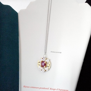 Könnte beinhalten: Halskette mit Silberkette und Anhänger. Der Anhänger hat eine goldfarbene Mitte mit einem großen, ovalen, roten Edelstein. Der Anhänger ist von einem weißen Filigranmuster umgeben. Der Text "jewelry by Et1762" ist sichtbar.