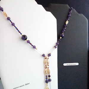 Peut inclure: Un long collier avec des perles violettes, des accents dorés et des tubes en verre transparent. Le collier a un design en forme de Y avec un pendentif. Le texte "Bijoux créateurs sautoir. Helmi" est visible.