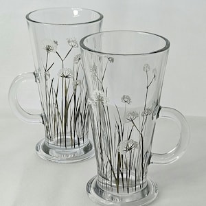 May include: Two clear glass mugs with a tapered shape, each featuring a handle and a sturdy base. The exterior of the glass is decorated with a delicate botanical print of white and dark green wildflowers and tall grass stems.