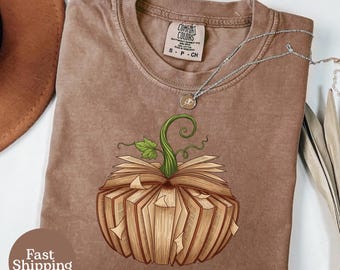 Könnte beinhalten: Braunes Comfort Colors T-Shirt mit einem Kürbisdesign aus offenen Büchern, grünem Stiel und Blättern. Eine silberne Halskette mit Anhänger ist auf dem Shirt drapiert. Das Shirt hat einen Rundhalsausschnitt und kurze Ärmel. Die Worte "Fast Shipping" stehen in einem braunen Kreis.