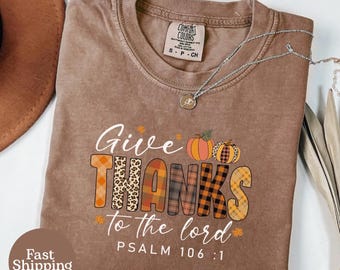 May include: Brown t-shirt with the text "Give Thanks to the lord" and "PSALM 106:1". The word "THANKS" is designed with a mix of patterns, including leopard print and plaid. Two pumpkins are above the word "THANKS". A silver necklace is on the shirt.