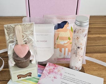 May include: A gift box with self-care items. Includes a chocolate spoon, clay mask, bath salts, and a card with text. The box is pink, and the items are arranged on a wooden surface.